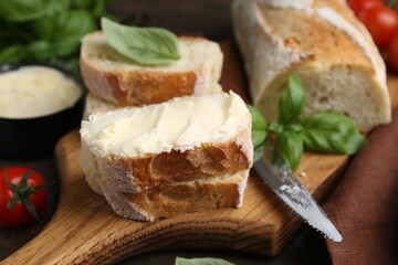 Slices of baguette with butter, basil, tomatoes and knife on wooden table, closeup