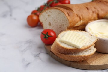 Cut baguette with butter and tomatoes on white marble table, closeup