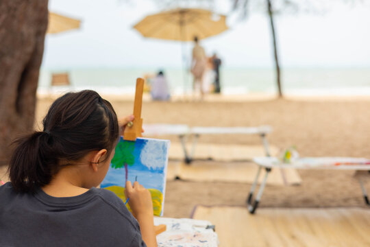 Over-the-shoulder view with blurred background of a young girl painting a seascape on an easel at beautiful beach, representing childhood creativity, outdoor learning, and a peaceful artist hobby. - Powered by Adobe