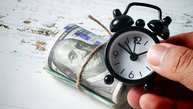 Financial Deadlines: Hand Holding Alarm Clock Next to Rolled US Dollar Bills