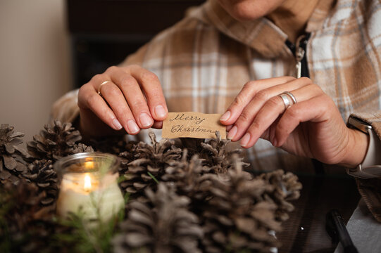 Hands Place A Merry Christmas Tag Among Pine Cones In Rustic Cozy Home Decor - Powered by Adobe