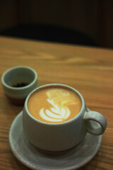 A close-up of a latte, featuring foam art in a speckled ceramic mug, sits on a wooden table. A small cup with dark liquid is positioned to the left, with the background heavily blurred.
