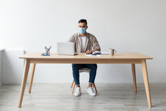 A young Middle Eastern businessman in a protective facemask types on his laptop at a home office. He is seated at a wooden desk with a notepad and a mug, focusing on his work during the pandemic.