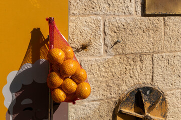 A mesh bag of bright oranges hangs against a vivid yellow wall with a graphic of an old person's face, beside a textured stone wall.