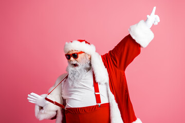 Santa in a red suit with white fur trim points up while wearing red suspenders and shades in a pink...