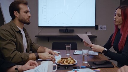 Professional office setting with coworkers sitting around a wooden table covered with business documents and some snacks as one colleague hands a financial report to a red-haired coworker - Powered by Adobe