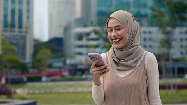 Portrait of Muslim female reads news about victory on smartphone stands against urban cityscape. Malaysian female in traditional headscarf wins in online lottery in modern city