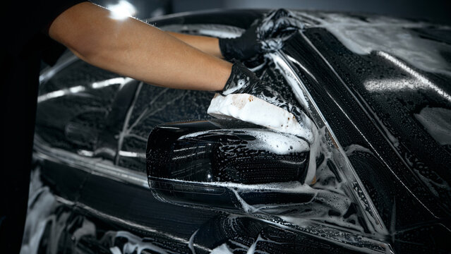 Hands washing car mirror with foam sponge during auto detailing. Concept of professional workflow visuals, precision cleaning, promotion for car care industry.