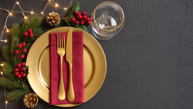 Christmas table setting with gold plate, red napkin, cutlery, glass, and festive decorations on dark background. Elegant holiday arrangement.