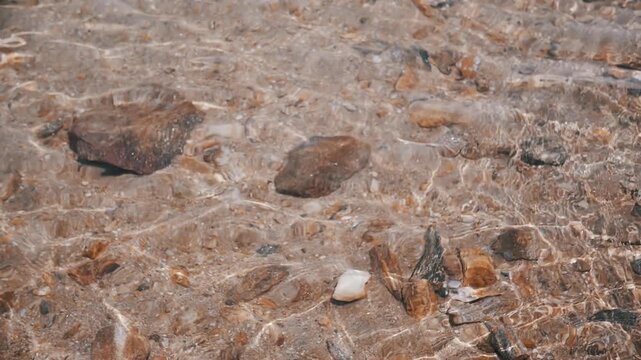 Underwater scene of a seabed where rocks and sand lie in clear sea water on a beach. Sun glare reflects on the surface of the water, swaying waves. Light pattern on sandy seabed. Abstract, texture.