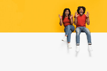 Fototapeta premium A happy couple shows excitement by sitting on a white surface. They wear matching red shirts and jeans, with big smiles and arms raised in celebration against a vibrant yellow background.