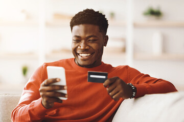 A young man sits on a couch, smiling while using his smartphone to shop online. He holds a credit card in one hand, enjoying the convenience of digital shopping in his cozy living room.