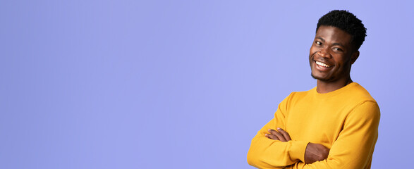 A young man stands confidently with his arms crossed, wearing a yellow sweater. He smiles broadly in front of a light purple background, giving off a cheerful and friendly vibe. © Prostock-studio