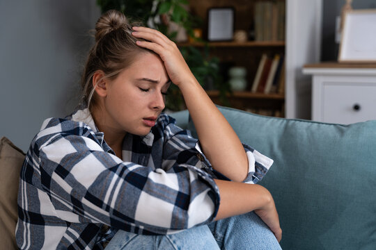 Young woman at home struggling with depression and anxiety, holding her head on sofa; signs of burnout, loneliness and overthinking, seeking health support and calm after stress, and sleepless nights