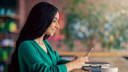 A young woman with long hair sits at a cafe, engaged with her smartphone. She wears a green top and appears focused. The background has soft lighting and greenery, creating a calm setting.