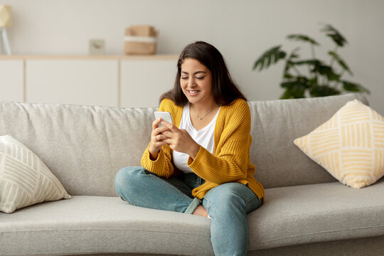 A cheerful young arab lady sits comfortably on a sofa at home, enjoying her smartphone while messaging friends or exploring new apps. She appears relaxed and engaged in her activity.