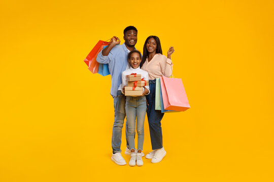 Three smiling black family members are joyfully posing together. A cheerful tween girl is holding a stack of wrapped gifts, while her parents carry colorful shopping bags.