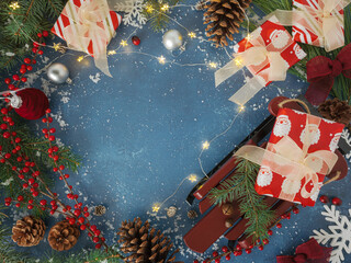 Overhead Christmas background with wrapped gifts and sled, festive velvet burgundy ornaments and bows, holiday twinkle lights and red berries, evergreen branches, pinecones on blue backdrop