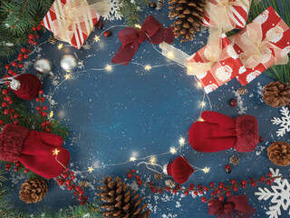 Overhead Christmas background with wrapped gifts and red gloves mittens, festive velvet burgundy ornaments and bows, holiday twinkle lights and red berries, evergreen branches, pinecones on blue