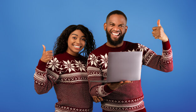 Xmas shopping online. Happy african american couple with laptop computer showing thumbs up, posing together in identical warm sweaters on blue background - Powered by Adobe