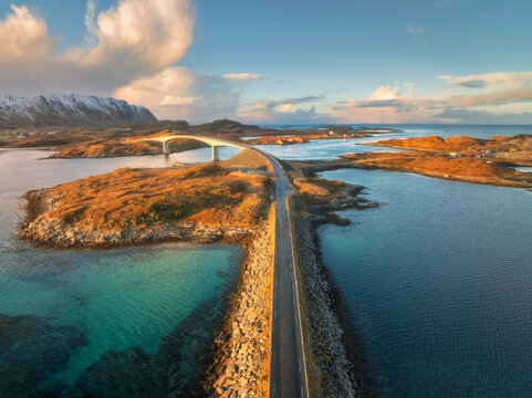 Aerial view of the Fredvang bridge and coastal road at sunset. Lofoten Islands, Norway. Turquoise sea, rocky islands, snowy mountains in warm evening light. Nordic landscape and Arctic road. Top view