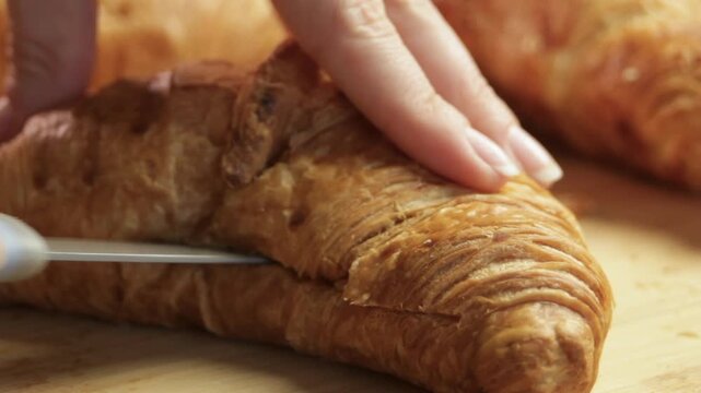 Female hands slice croissant on wooden board, showing croissant cut and croissant texture. Knife moves through flaky pastry. Concept of croissant as base for sandwich in French breakfast.