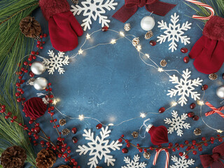 Overhead Christmas flatlay with red knit gloves mittens and snowflakes, holiday red velvet and silver ornaments, berries, twinkle fairy lights, festive pinecones, candy canes on blue backdrop. 