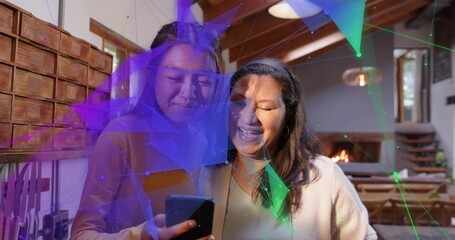 Holding smartphone, woman on left showing screen to woman in cardigan at home, with digital overlay