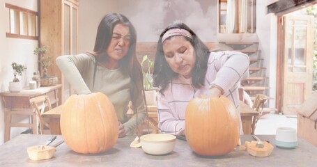 Carving two women leaning over table in cozy kitchen wearing headband and using pumpkins and tools