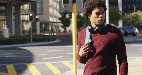Walking man gripping light blue backpack strap at crosswalk, maroon sweater, headphones, copy space
