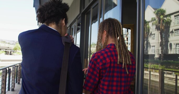 Walking two men, navy blazer, plaid shirt, on boardwalk with bag strap and glass reflections