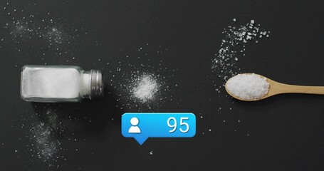 Showing tipped glass shaker spilling salt beside wooden spoon on dark matte tabletop, blue 95 badge