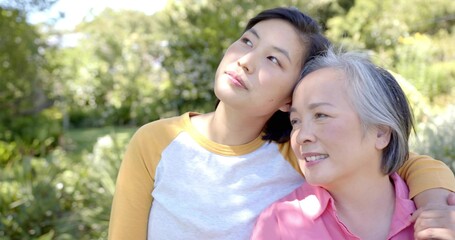 Leaning mother and daughter touching heads in sunlit park, wearing pink blouse and mustard sleeves