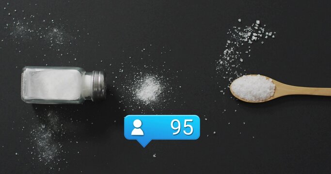 Showing tipped glass shaker spilling salt beside wooden spoon on dark matte tabletop, blue 95 badge - Powered by Adobe