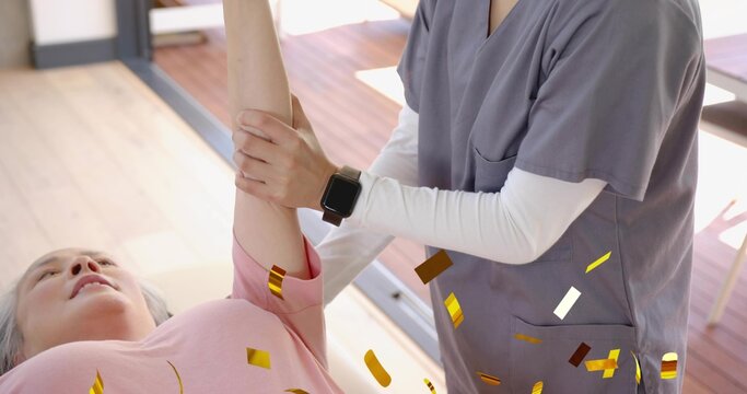 Caregiver in gray scrubs holding senior woman wearing pink top on table in therapy room, smartwatch - Powered by Adobe