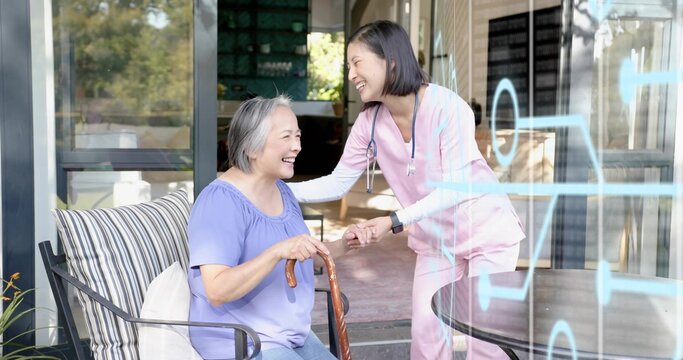 Smiling senior Asian woman holding cane on patio, pink-scrubbed caregiver with stethoscope leaning
