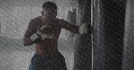 Striking shirtless boxer punching heavy bag at gym, wearing dark shorts, white wraps and gloves