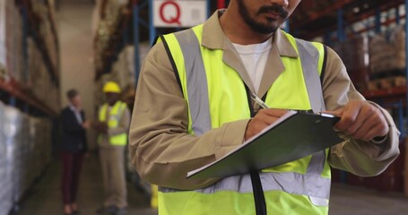 Writing mid adult warehouse worker wearing hi-vis vest tan shirt holding clipboard in aisle