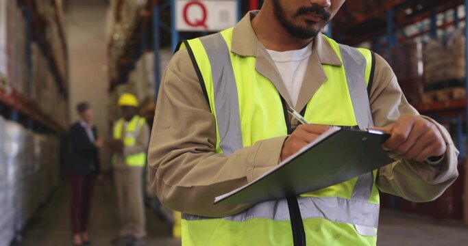 Writing mid adult warehouse worker wearing hi-vis vest tan shirt holding clipboard in aisle - Powered by Adobe