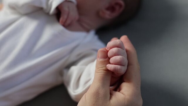A newborn in a white bodysuit against a gray background. The baby is resting with its legs spread out. The mother holds the baby's tiny feet in her hands. The girl strokes the doll's toes