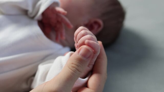 A newborn in a white bodysuit against a gray background. The baby is resting with its legs spread out. The mother holds the baby's tiny feet in her hands. The girl strokes the doll's toes