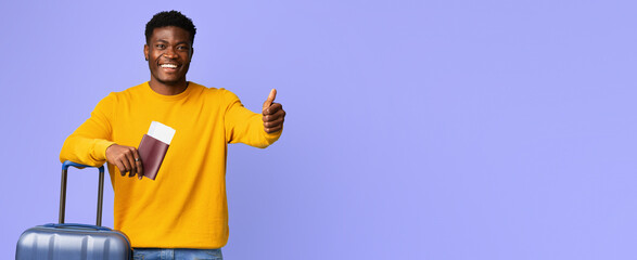 A young man is smiling and giving a thumbs up while standing next to a suitcase. He holds a passport in one hand, expressing anticipation for his upcoming journey.