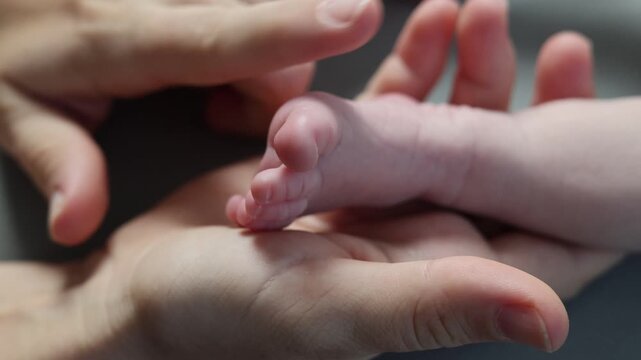 A newborn in a white bodysuit against a gray background. The baby is resting with its legs spread out. The mother holds the baby's tiny feet in her hands. The girl strokes the doll's toes.