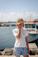 Fototapeta premium Girl standing on a pier near boats and yachts, wearing a hat and summer outfit. Relaxed vacation atmosphere by the marina under a bright blue sky. 
