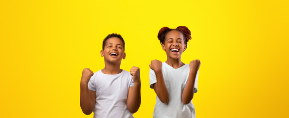 Two children stand side by side, smiling widely with their fists raised in celebration. Their excitement fills the air against a cheerful yellow backdrop, capturing a moment of pure joy and happiness.