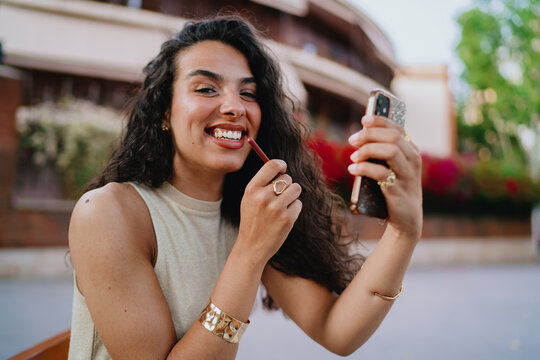 Confident woman laughs mid-application of lip liner, blending beauty habits with interactive smartphone use in a connected lifestyle.