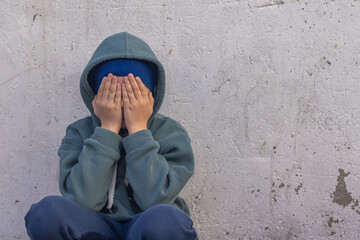 A little boy sitting against the white wall, covering his face with his hands, looking upset