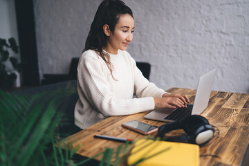 Confident freelancer smiling at laptop while typing, immersed in digital workflow, showing ease with technology and mastery of flexible modern working culture and virtual presence.