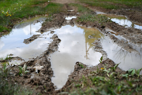 Rural dirt road with large puddles after rainfall and tire tracks. Muddy countryside path reflecting cloudy sky, symbol of weather, travel or rustic lifestyle.