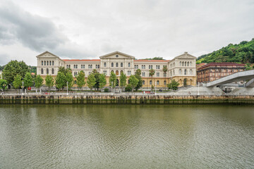 A view of a large stone white building on the embankment of the Nervion River in the city of Bilbao, Spain. The picture was taken on a cloudy day with the sky covered in clouds